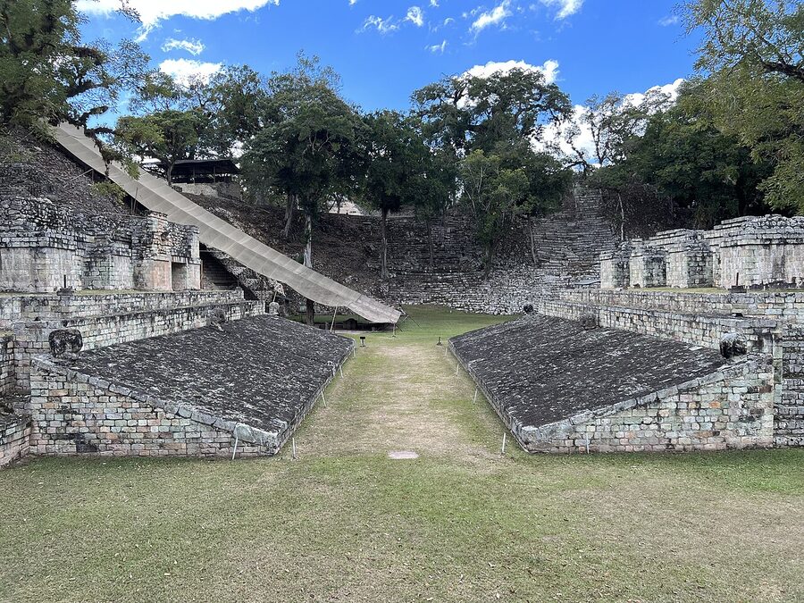 The Ball Court at Copán with sloping stone aprons on both sides and a grassy alley between, surrounded by trees and a low pyramid in the background