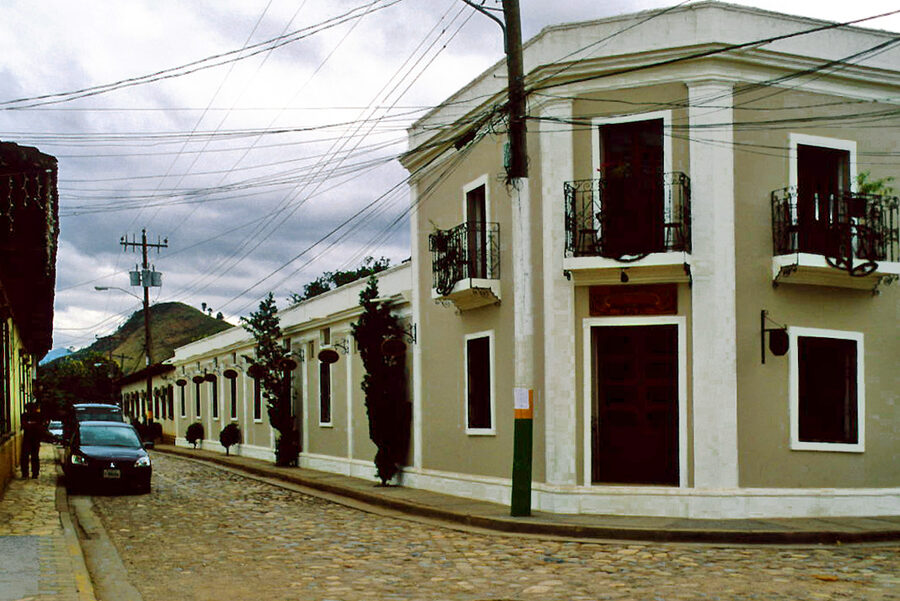 A cobblestone street corner in the town of Copán Ruinas, with whitewashed colonial buildings and small wrought-iron balconies under a grey sky